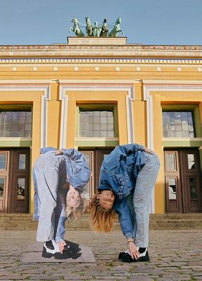 A photo of a model mirroring a cardboard cutout. Both touching their toes wearing jeans and trucker jack from the Levi's x Ganni collection.
