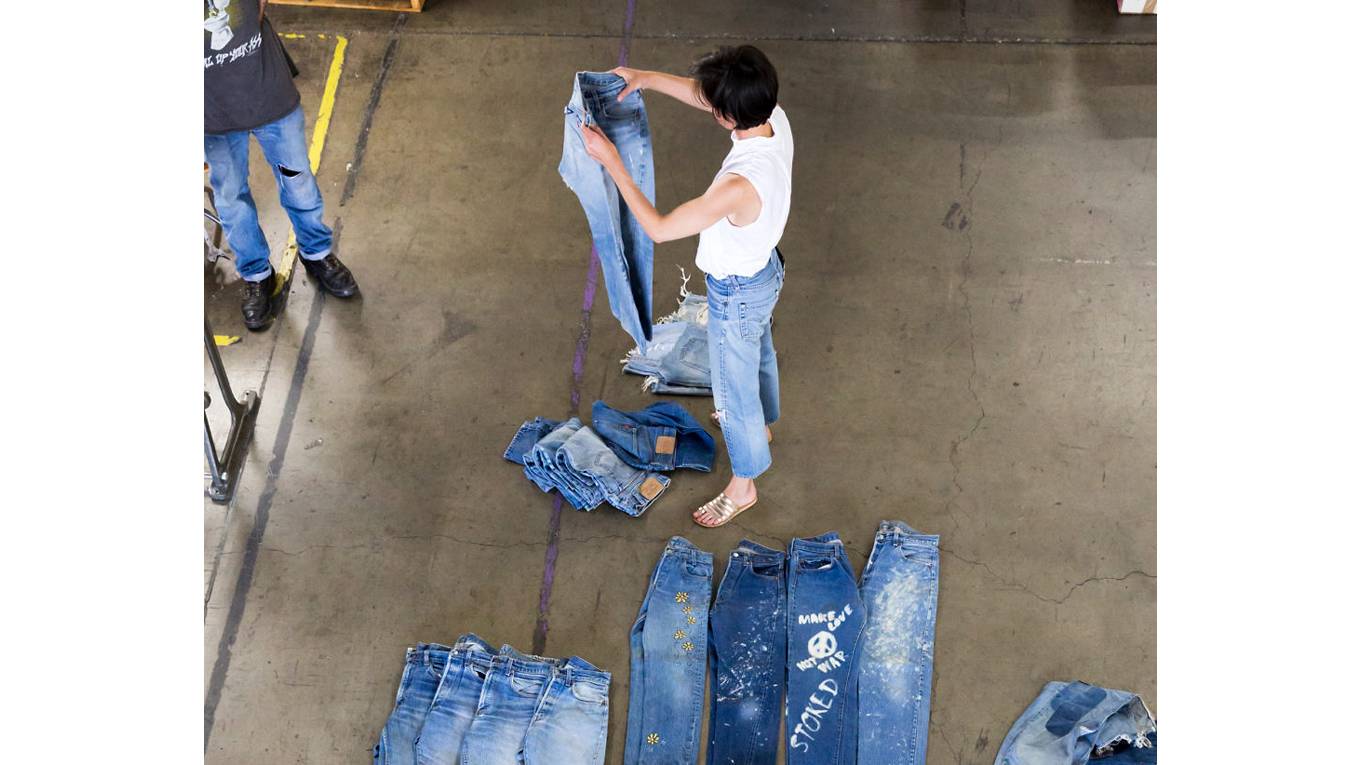 Person holding a pair of jeans up and a bunch of jeans laying on the ground around.