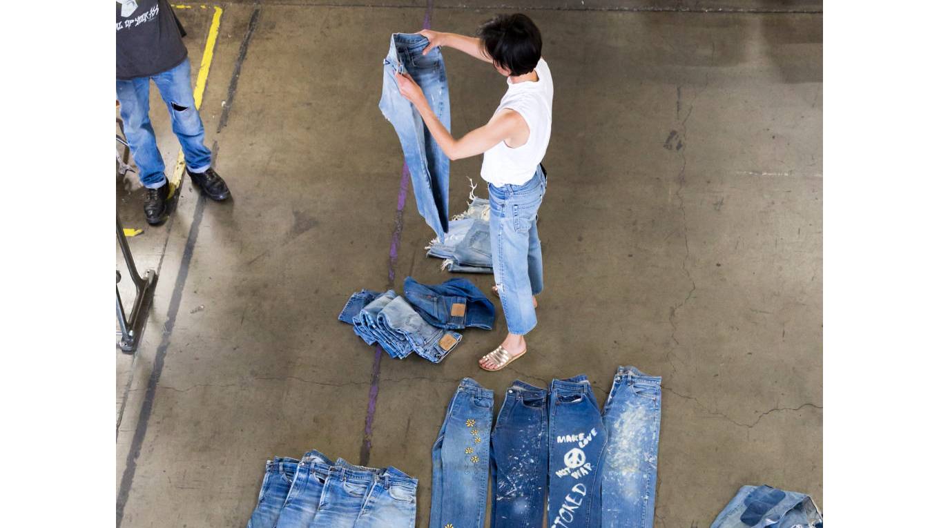 Person holding a pair of jeans up and a bunch of jeans laying on the ground around.