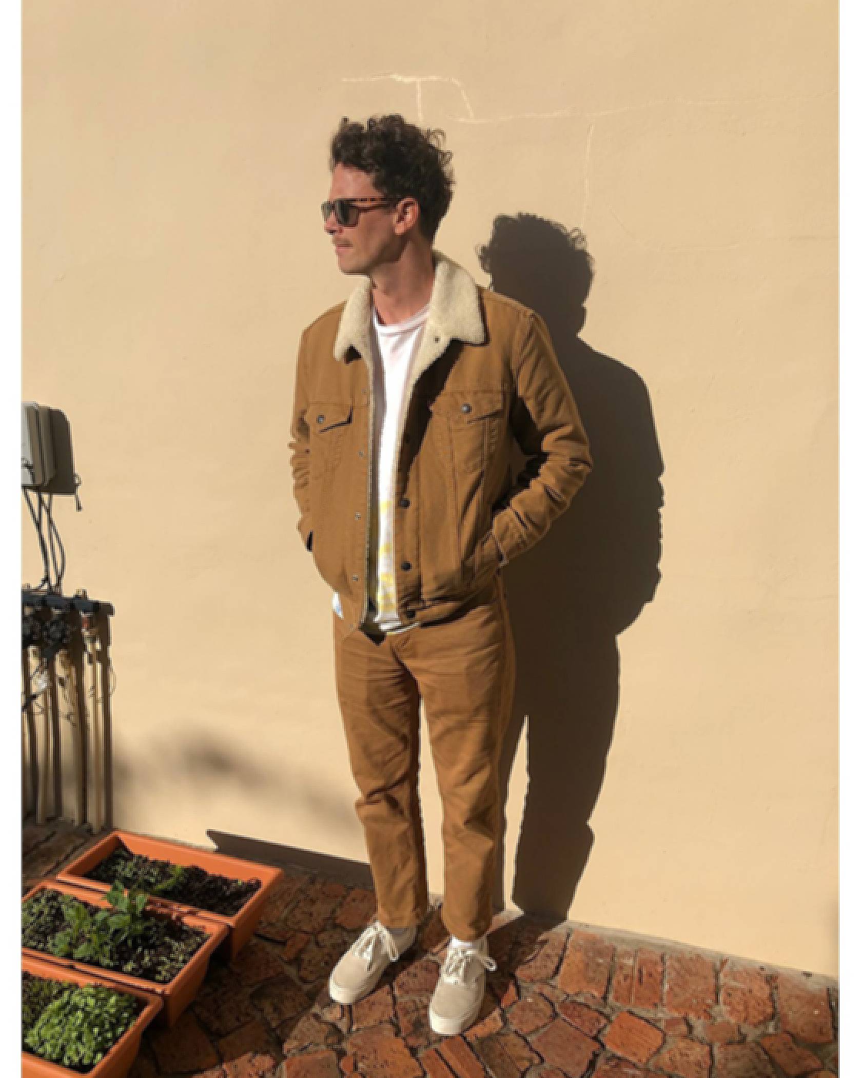 Photo of David Davey standing against a cream wall outside. He's wearing a brown corduroy jacket, a white tee, and tan 514s with tan sneakers.