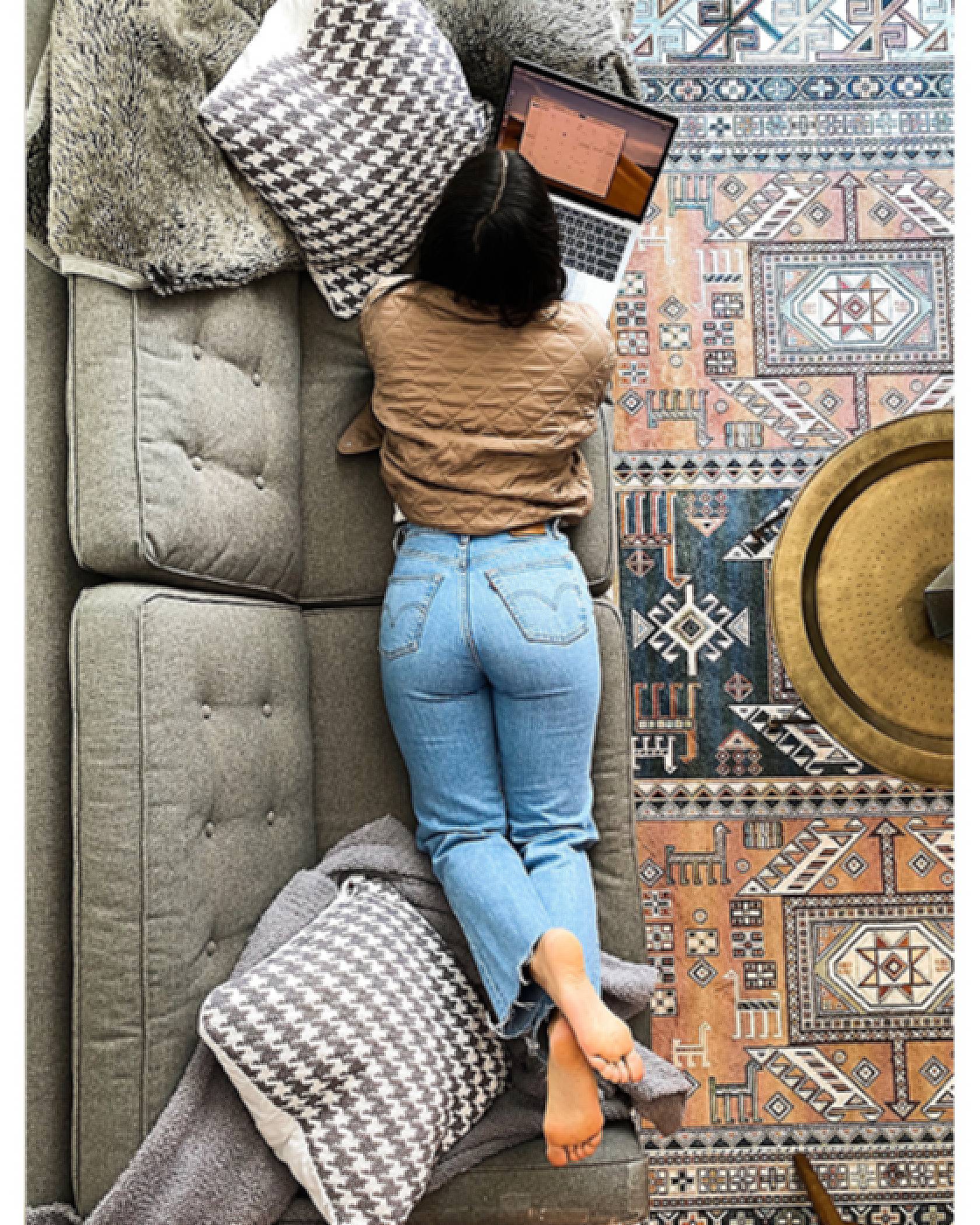 Aerial shot of Zoe Chrysochoos lying on her couch on her stomach with her laptop open. She is wearing a brown top and Levi's jeans.