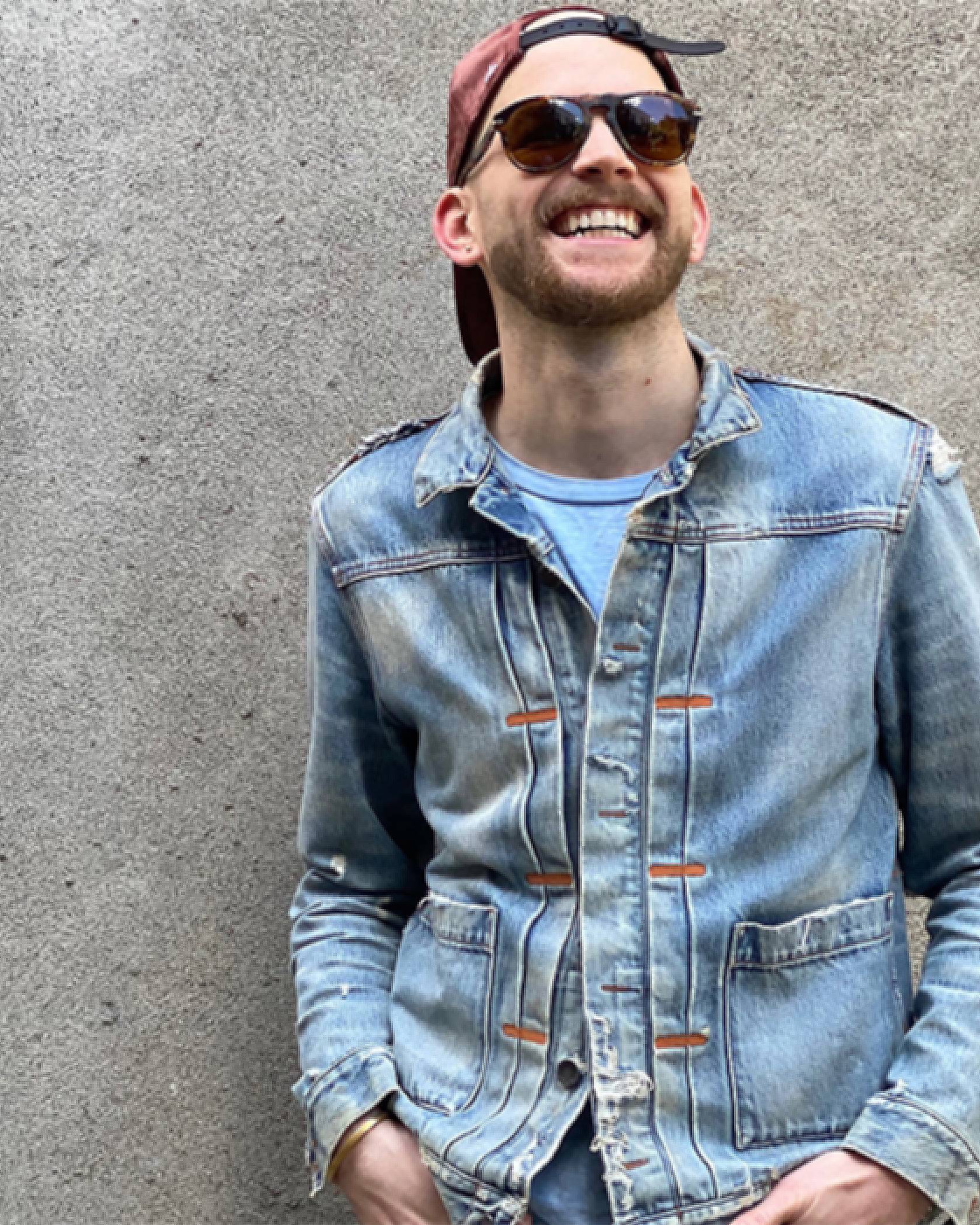 Photo of Austin McCune standing against a concrete wall. He is wearing a baseball hat backwards, sunglasses, and a faded Levi's Trucker Jacket.