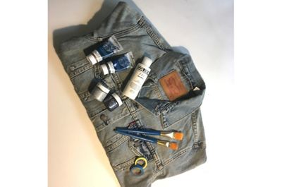 Image of a folded blue denim trucker jacket with paint, paint brushes and neon colored tape laying on top.