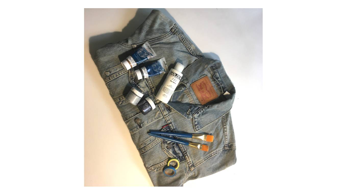 Image of a folded blue denim trucker jacket with paint, paint brushes and neon colored tape laying on top.