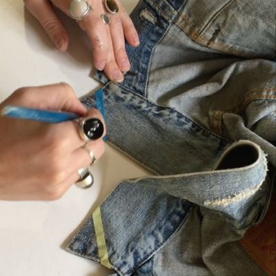 Image of the edges of a blue trucker jacket and a hand with multiple rings on it using a paintbrush to paint glitter on the edges of the jacket.