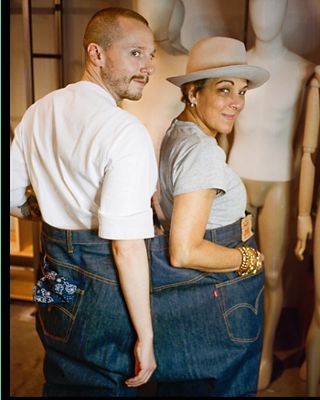 Photo of Ezra and Bettina standing in the same pair of oversized Levi's jeans, one person per leg. Ezra is wearing a white tee and Bettina is wearing a grey tee with a cream hat and gold bracelets on her right wrist.