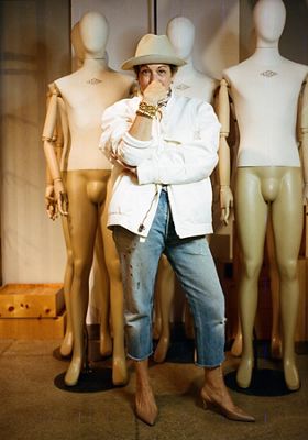 Photo of Bettina Sapien wearing a white bomber jacket and cropped Levi's blue jeans with tan slingback heels. Her arms are crossed and there is a stack of gold bracelets on her right wrist and she has on a cream hat.