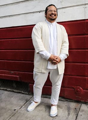 Photo of Gem standing outside in front of a red and white wall. He is wearing a white button up, a cream jacket, and white bottoms with cream sneakers.