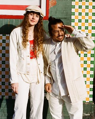 Photo of Haley and Gem standing in front of a green and yellow wall in San Francisco. They are both wearing white head-to-toe.