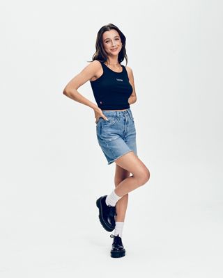 Emma Chamberlain standing in a tank top and jean shorts smiling.