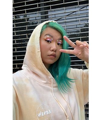 A photo of Mi-Anne wearing a Levi's sweatshirt with the hood up looking at the camera with a peace sign.