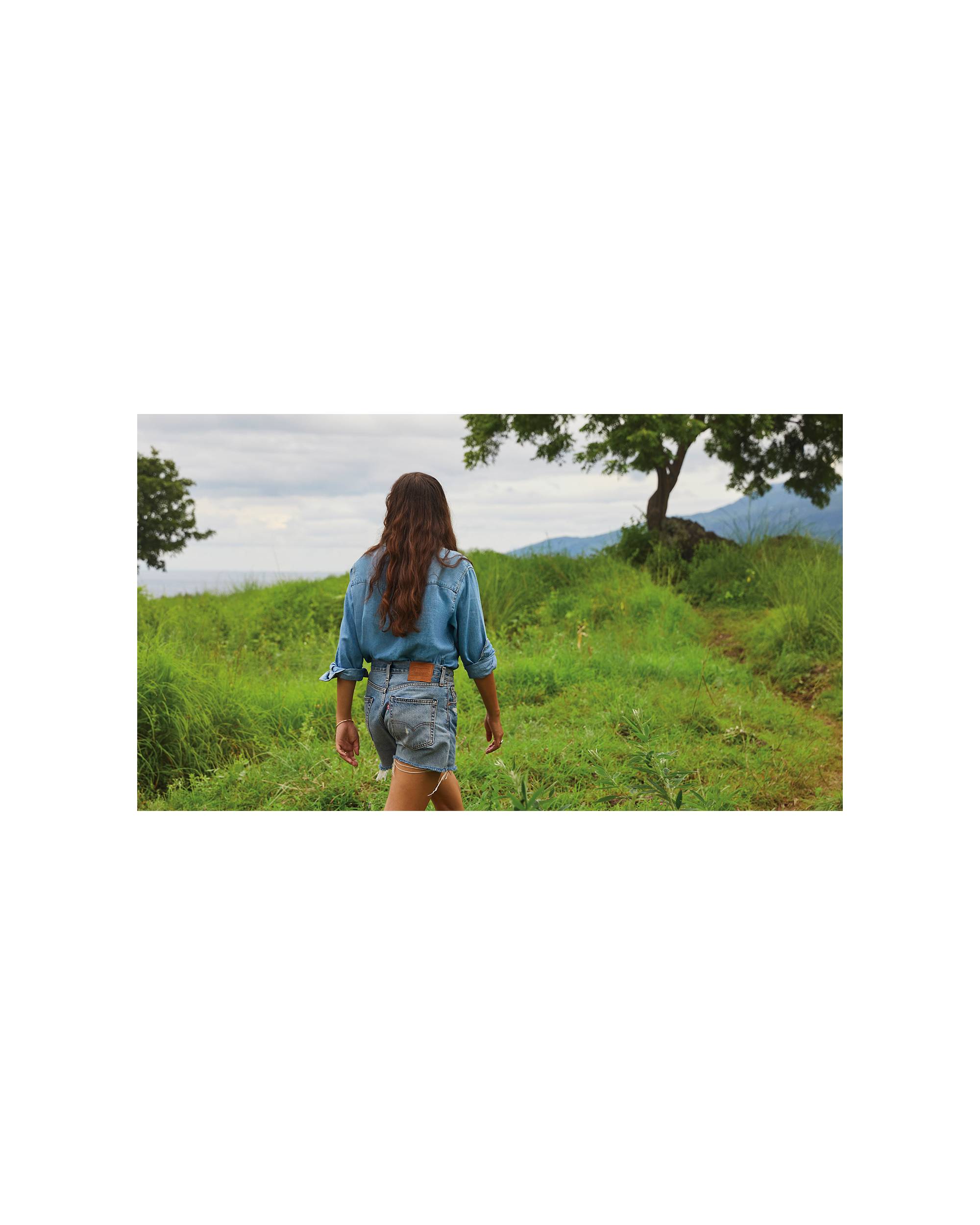 A photo showing Melati from behind walking in a lush green field wearing a Levi's button up and Levi shorts