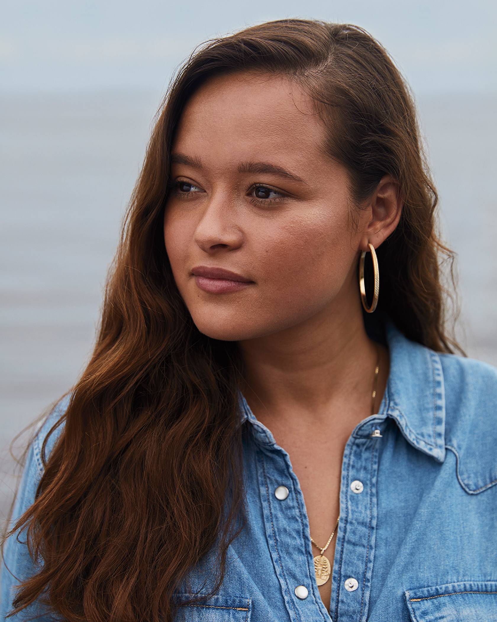 A portrait shot of Melati wearing a Levi's button up shirt, a gold circle necklace and gold hoop earrings