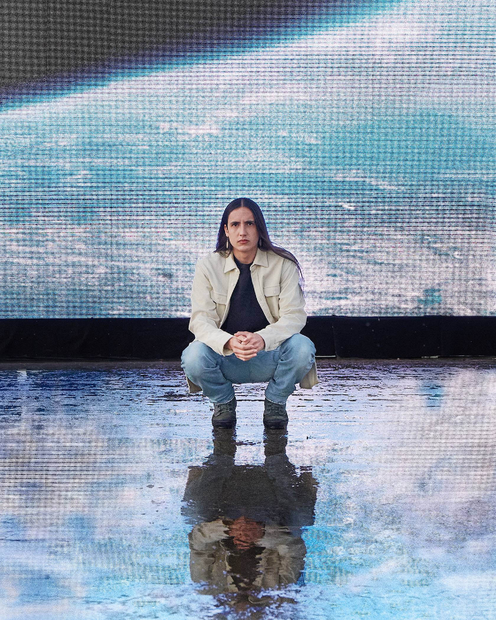 Xiuhtezcatl squatting in front of a screen wearing Levi jeans and a button up shirt.