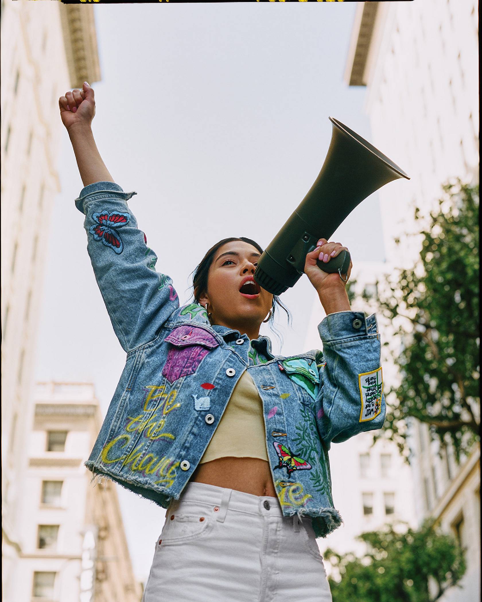 A photo of Xiye standing with a loud speaker and her fist raised wearing white Levi jeans and a custom Levi's trucker jacket that has the words, "Be the Change" written on it.