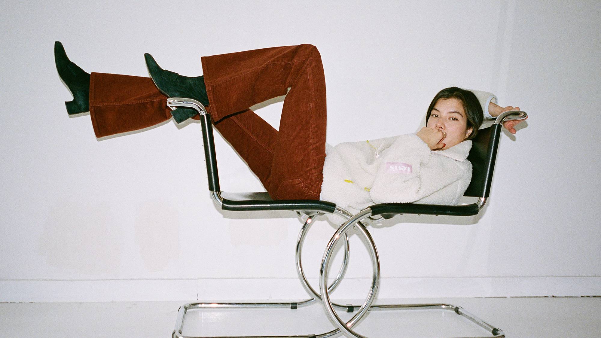 A women laying across two chairs with her feet on the back wearing a cream colored sweatshirt and rust colored 70's styled pants