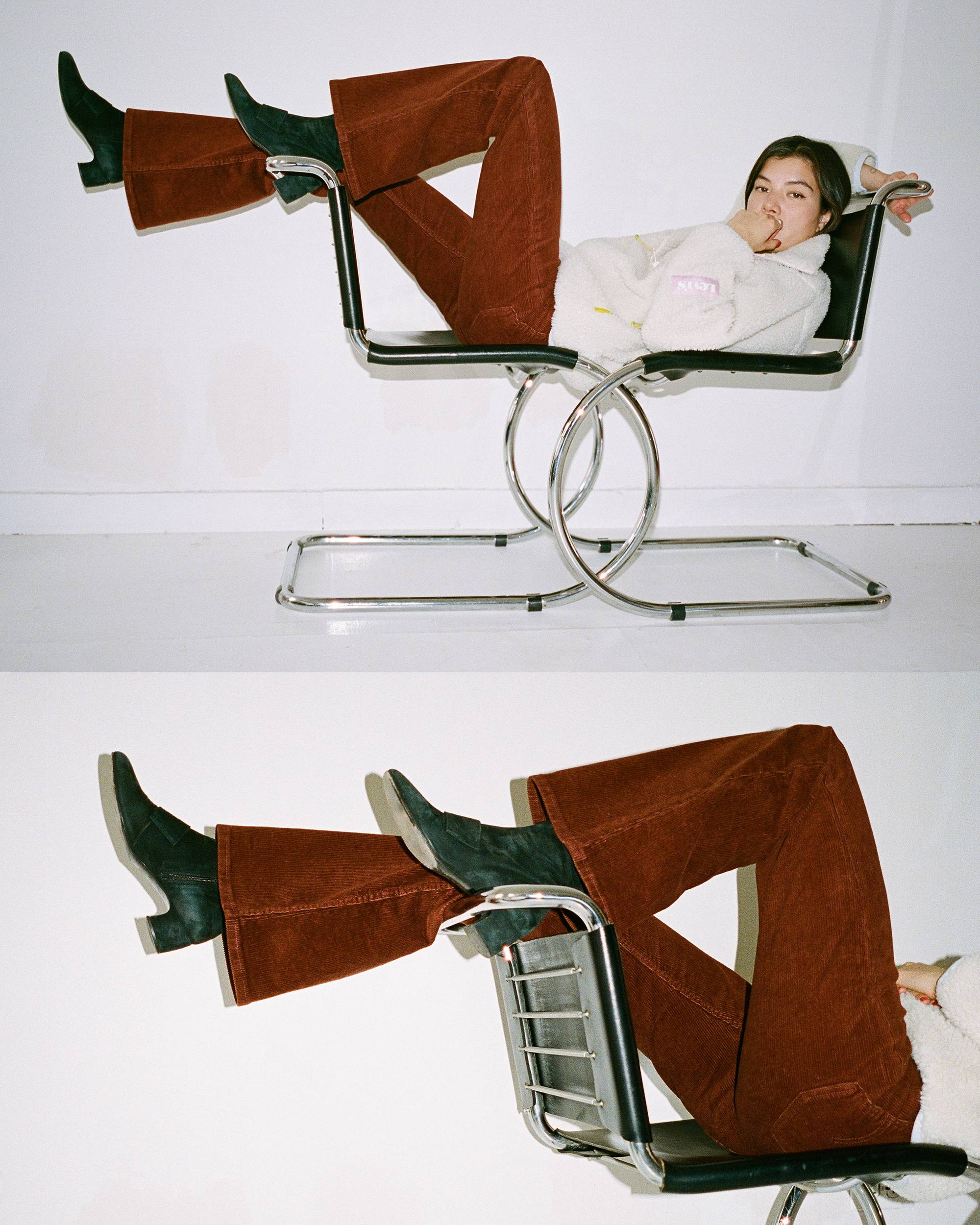 A women laying across two chairs with her feet on the back wearing a cream colored sweatshirt and rust colored 70's styled pants
