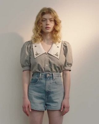 MP4 of a woman standing in front of a white wall with a black checkered button up.