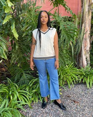 Woman wearing a sweater vest and denim jeans standing in front of green bushes.