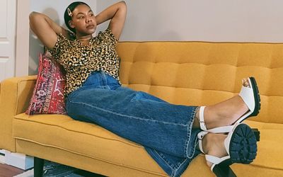 Photo of Natasia Berry sitting on a mustard yellow couch. She is wearing a leopard print shirt, Levi's jeans, and white heels.