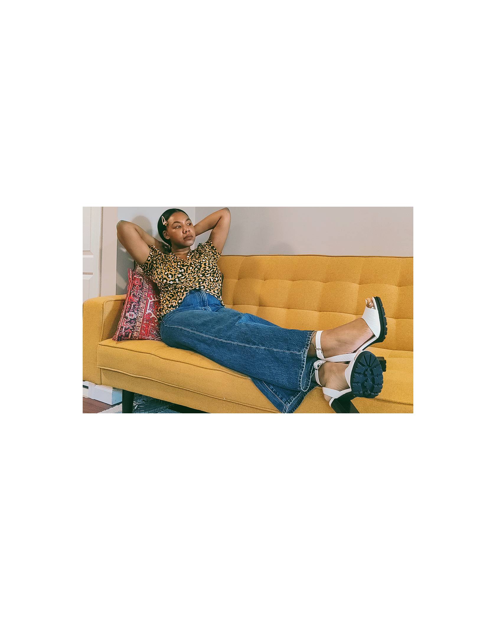 Photo of Natasia Berry sitting on a mustard yellow couch. She is wearing a leopard print shirt, Levi's jeans, and white heels.