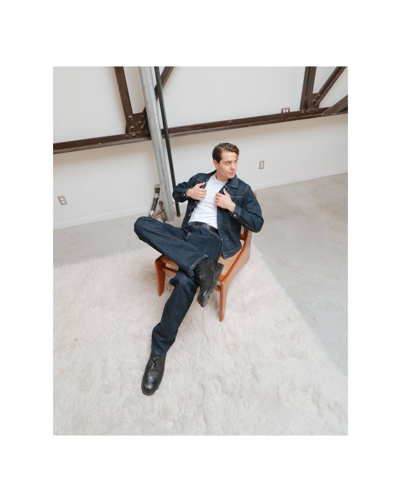 Gabe sitting on a wooden chair in an industrial room wearing jeans, matching jacket and white tee