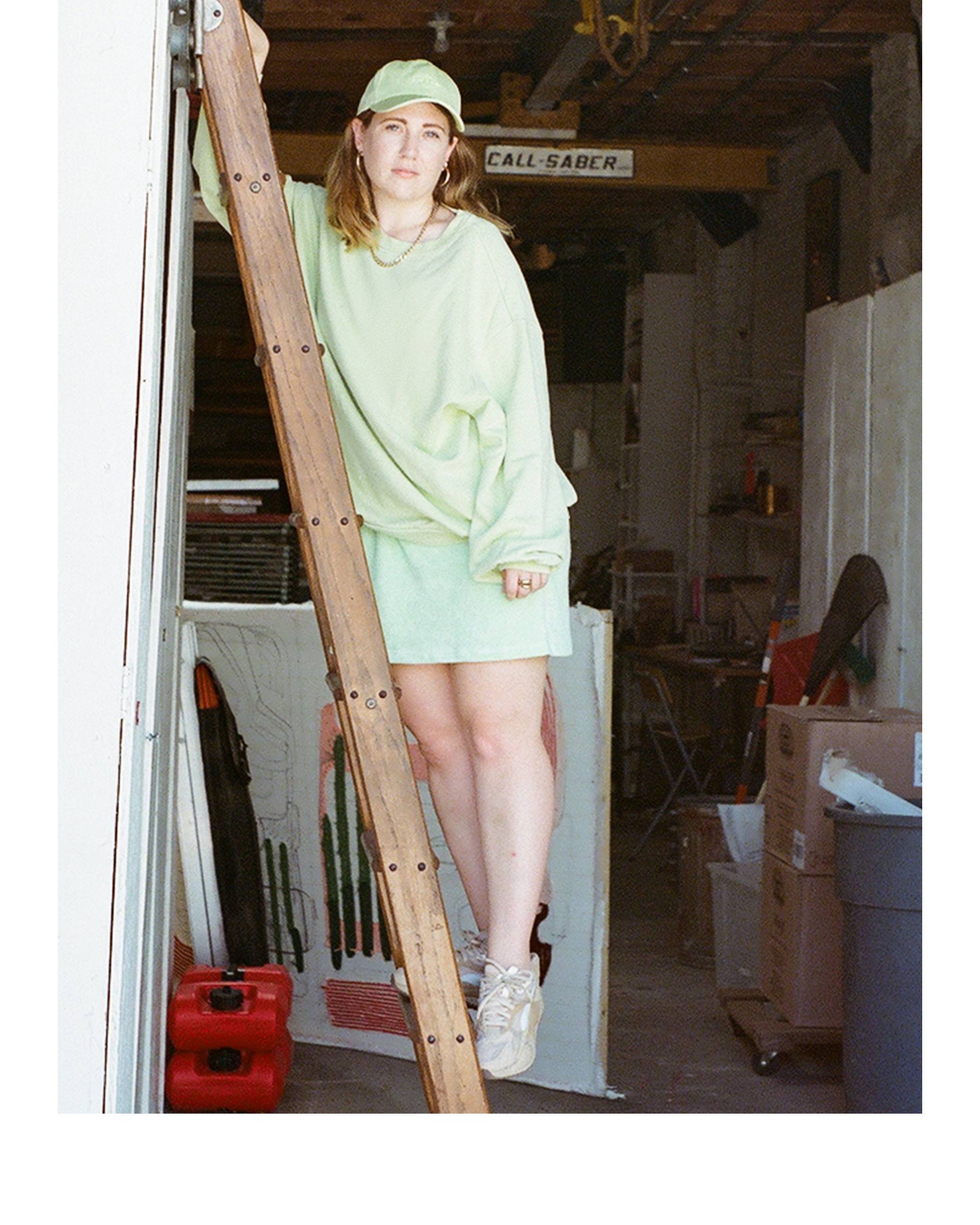 Caroline Kaufman wearing a levis outfit standing on a ladder in a garage with art in the background