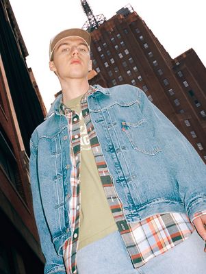 Model wearing denim jacket and plaid shirt layered over green tshirt and blue jeans with a city skyline behind him