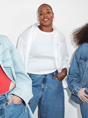 Model wearing white tank with white button down layered on top and blue jeans with two other models leaning off frame