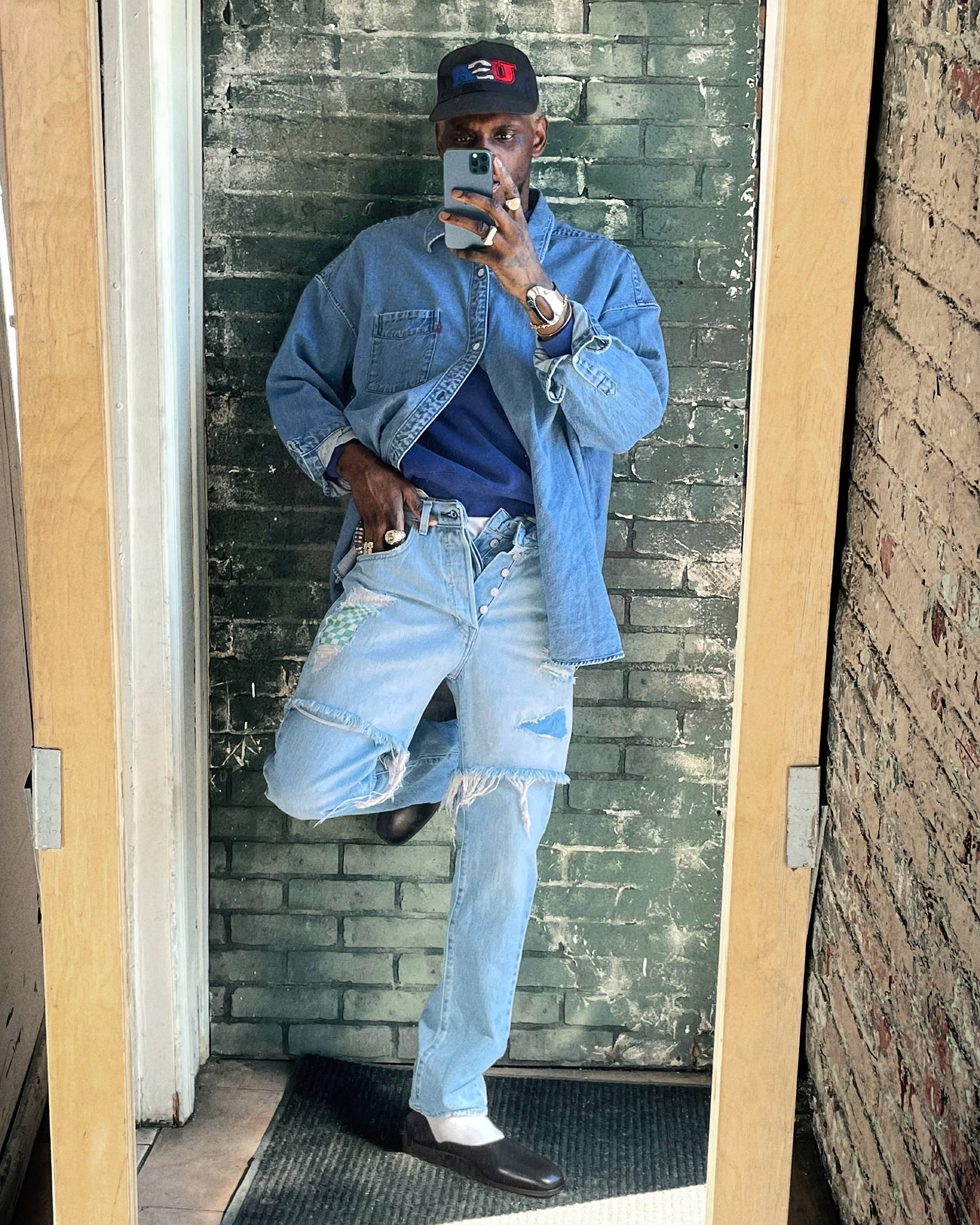 Image of man leaning against wall taking photo of his styled denim look