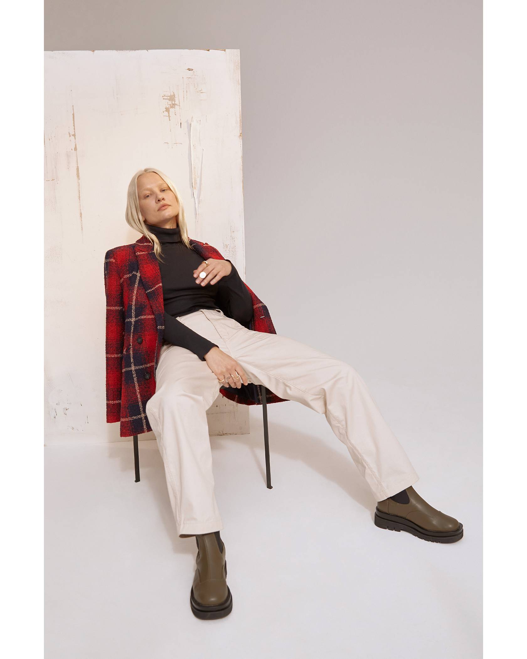 Model wearing red plaid jacket, black turtleneck, cream pants, and green boots