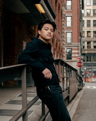 Lead Tailor Miguel Adrian Corachea leaning against a railing with brick buildings in the background while wearing a dark wash Levi's® Trucker Jacket and dark wash Levi's® jeans.