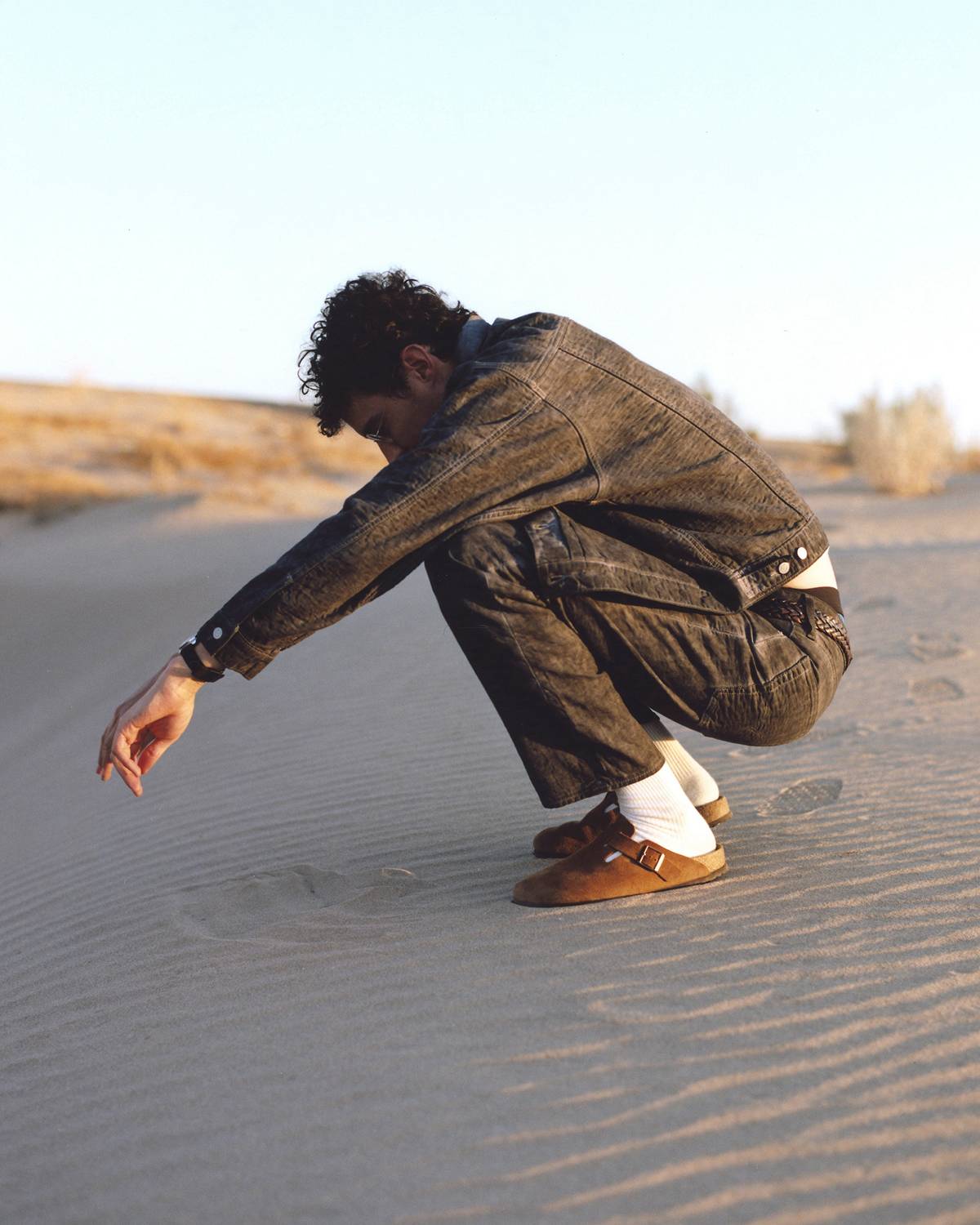 Image of male model with short, curly brown hair, crouching on sand, wearing the Stüssy & Levi's® Season 2 trucker jacket and jeans.