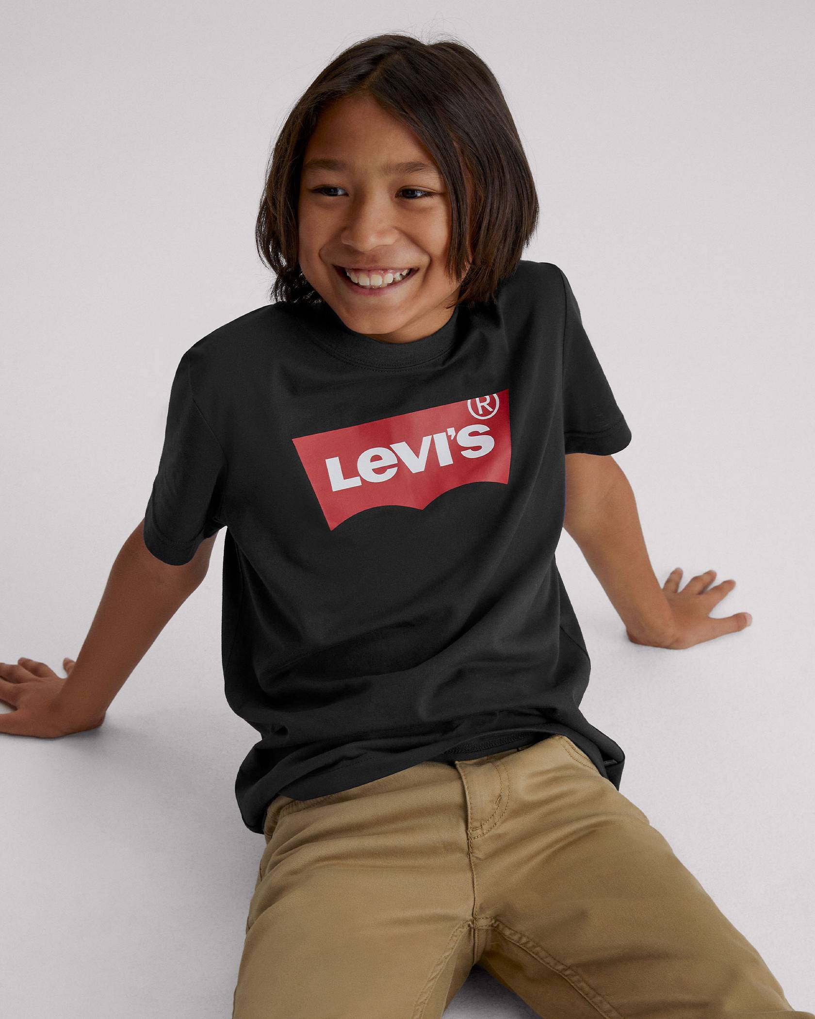 Model sitting on the ground in kids' khaki pants and black logo batwing tee