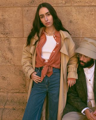 Three images of Levi's® employees Adeline Hocine and Paul Bellas. The left image is of Paul in a green camo jacket. The middle image is of Adeline in an orange blouse and camel trench coat with her arm resting on Paul sitting against a wall. The right image is of Adeline sunkissed while sitting in a pair of blue jeans and surrounded by monstera plants.