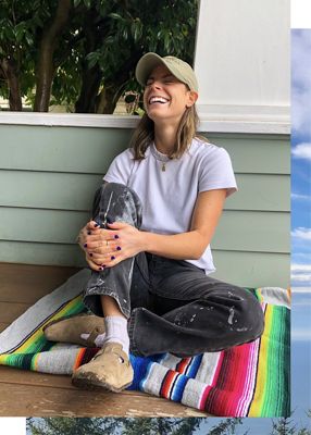 Photo of Lauren Bigelow sitting on a blanket on her porch. She is wearing a tan baseball cap, white tee, and paint-splatter black Levi's jeans with white socks and Birkenstocks. She is laughing.