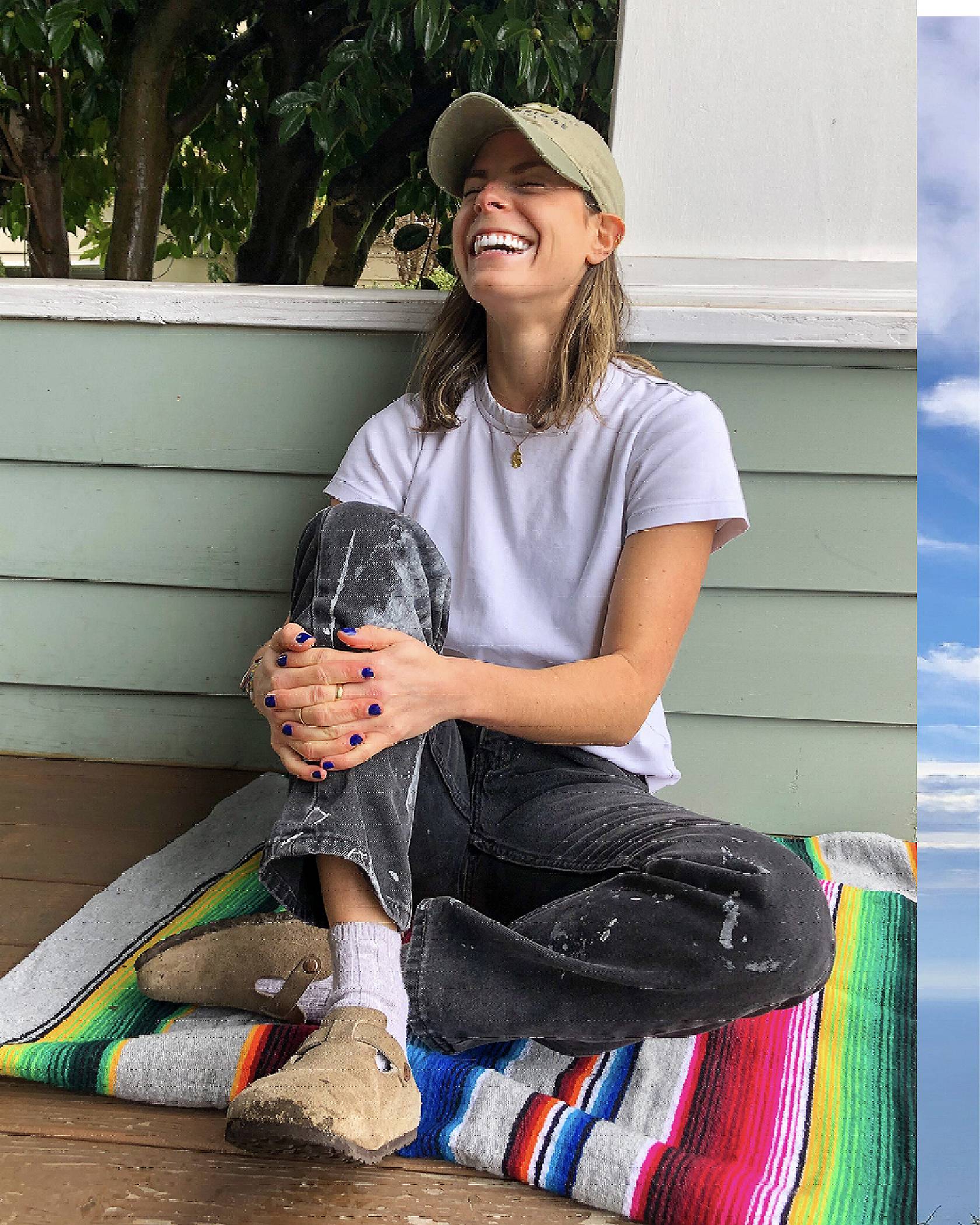 Photo of Lauren Bigelow sitting on a blanket on her porch. She is wearing a tan baseball cap, white tee, and paint-splatter black Levi's jeans with white socks and Birkenstocks. She is laughing.