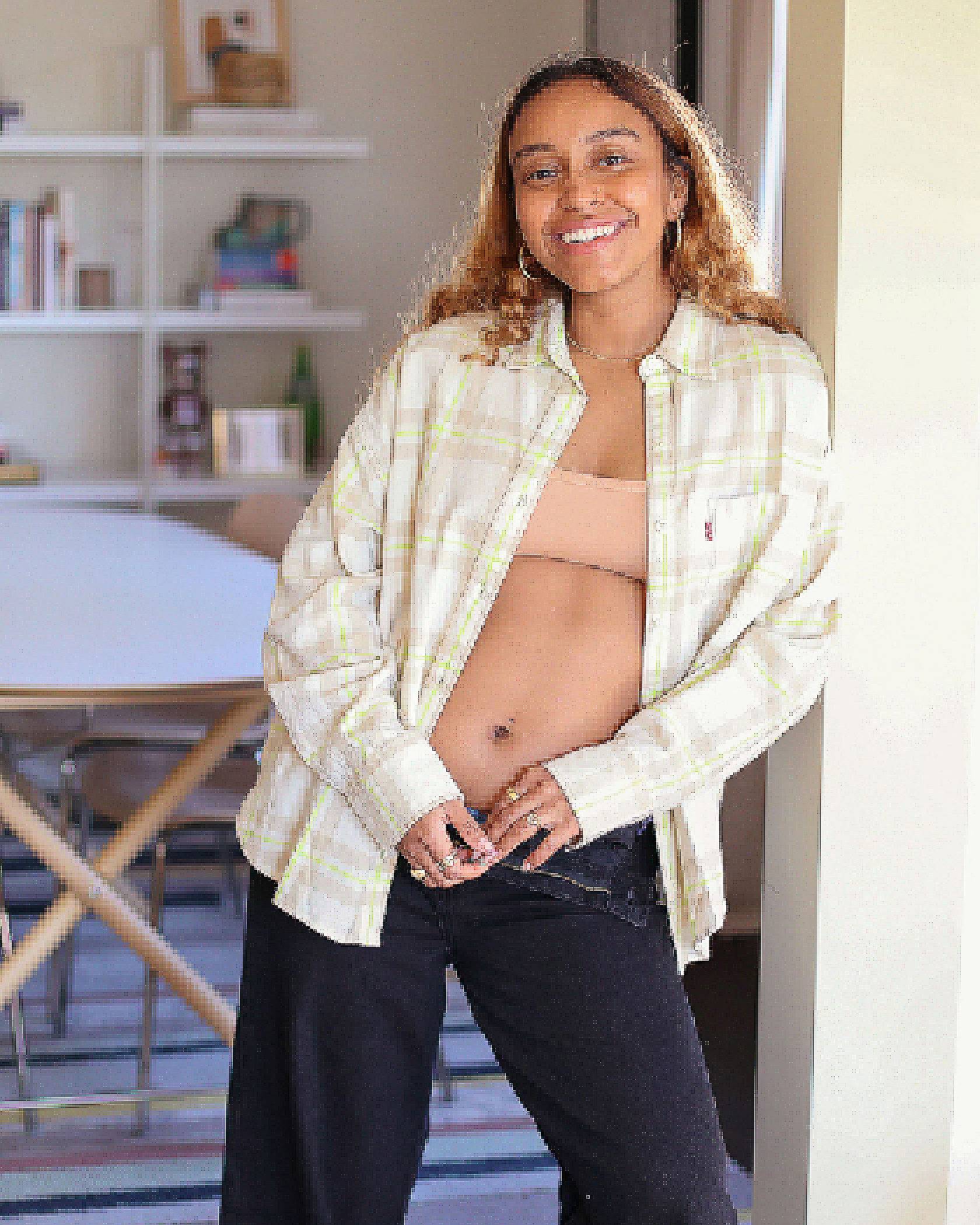 Portrait of Evelynn Escobar-Thomas standing in a doorway inside her home smiling. She is wearing a peach sports bra, a cream and yellow flannel, and black jeans.