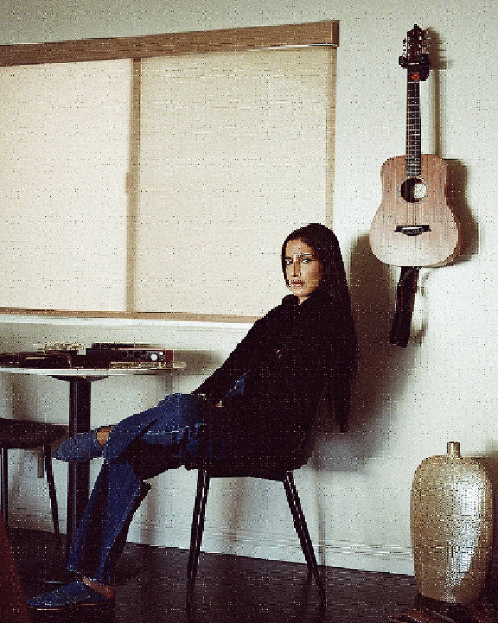 Portrait of ABIR sitting in a chair with a guitar hanging on the wall behind her.