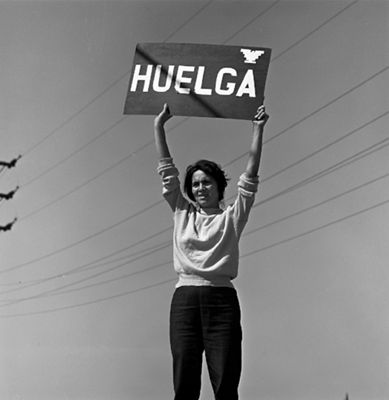 A black and white photo of Dolores standing holding a sign up that reads, "Huelga"