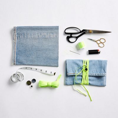 Close up image of all the things needed for the DIY laid out on a white background, including: a pair of old jeans, flat elastic cord, measuring tape, scissors, a button, sewing needle, sewing pins, thread and a lighter.