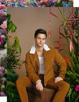 Brandon Flynn sitting on a wooden stool, surrounded by plants.
