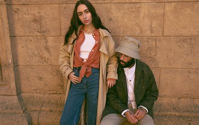 Image of Adeline Hocine and Paul Bellas standing against a brown wall. Adeline is wearing a tan colored trenchcoat, orange blouse, white tank top and blue denim jeans while Paul is wearing a tan colored bucket hat, green camo jacket, white button down shirt and tan colored cargo pants.