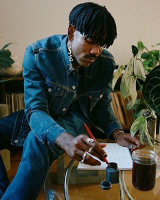 Photo of Jerrod La Rue in a Levi's Trucker Jacket and jeans writing in his notebook with a glass of iced coffee on the table.