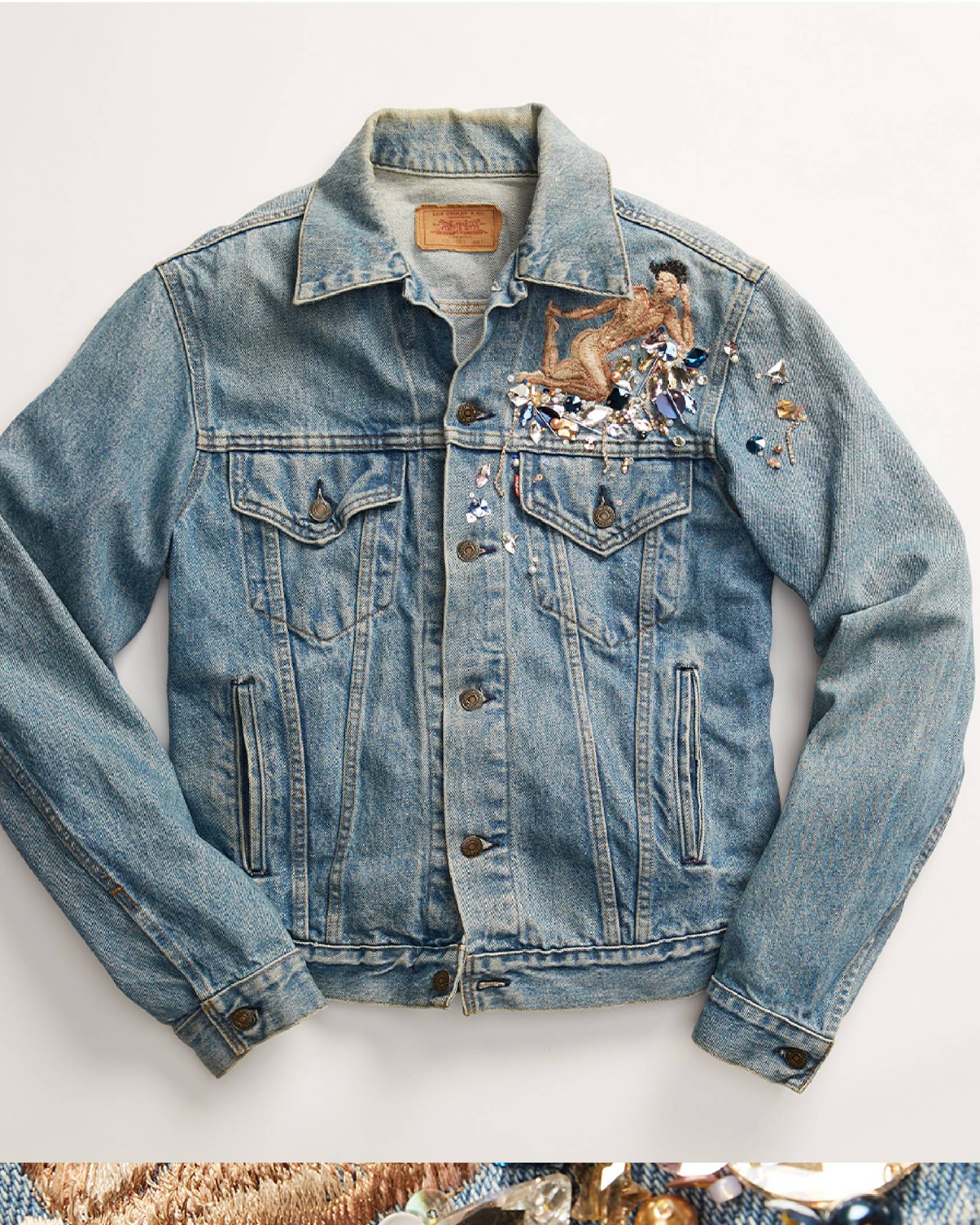 Laydown image of a light wash trucker jacket embroidered with a person sitting on top of a bed of jewels on the upper left side of the front of the jacket, above the trucker jacket's pocket.