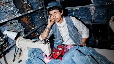 Jimmy Amarez posing in front of a wall of jeans using a sewing machine.