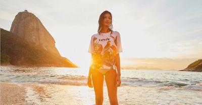 A woman standing on the beach at sunset wearing a Levi white T shirt with jungle animals and plant print and a bikini bottom.