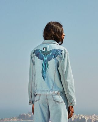 A woman facing away from the camera wearing a Levi's jean jacket with a blue parrot across the shoulder blades and Levi jeans.