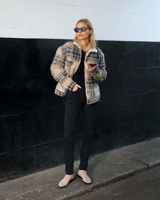 Photo of Claire Rose standing in front of black and white wall wearing tan plaid print trucker, black shirt, black jeans, and tan boots