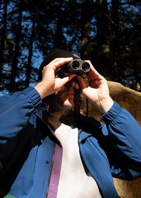 Video flipping through three images of a man and a woman in the woods. In one of the images, the woman is wearing a green sweatshirt and black denim jeans while holding a dog leash. One of the other images shows a man on top of a pair of large rocks, wearing a grey sweatshirt, black beanie and looking to the side. The last image includes the same man wearing a blue jacket and black beanie while looking through a pair of binoculars.
